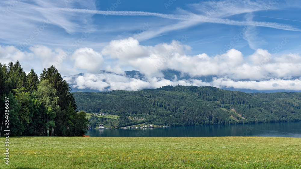 green summer meadow on the edge of a wood with view to lake Millstatt, in the background beautiful alpine landscape with some houses, mountain and forest, blue sky and white clouds, Carinthia, Austria green summer meadow on the edge of a wood with view to lake Millstatt, in the background beautiful alpine landscape with some houses, mountain and forest, blue sky and white clouds, Carinthia, Austria