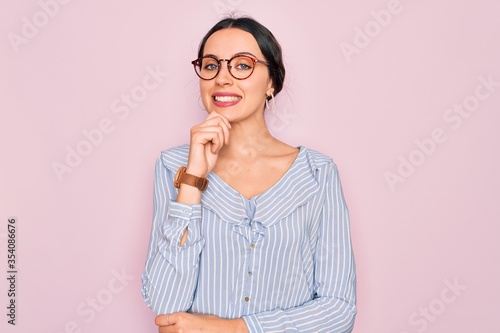 Young beautiful woman wearing casual striped shirt and glasses over pink background looking confident at the camera with smile with crossed arms and hand raised on chin. Thinking positive.