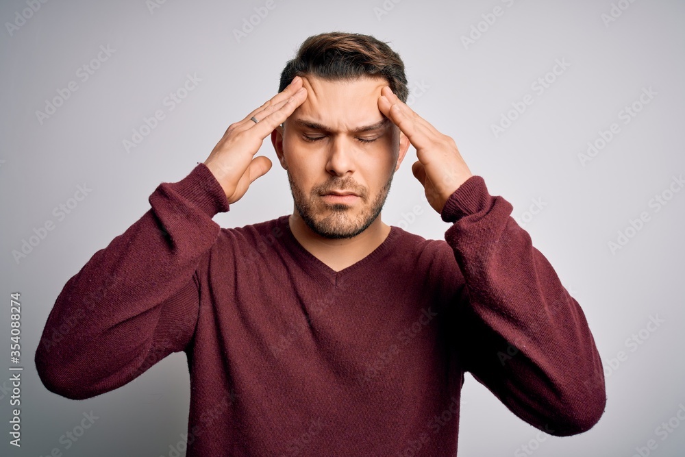 Young man with blue eyes wearing casual sweater standing over isolated background suffering from headache desperate and stressed because pain and migraine. Hands on head. Young man with blue eyes wearing casual sweater standing over isolated background suffering from headache desperate and stressed because pain and migraine. Hands on head.