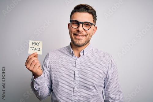 Young business man with blue eyes holding pay taxes word on paper note with a happy face standing and smiling with a confident smile showing teeth © Krakenimages.com