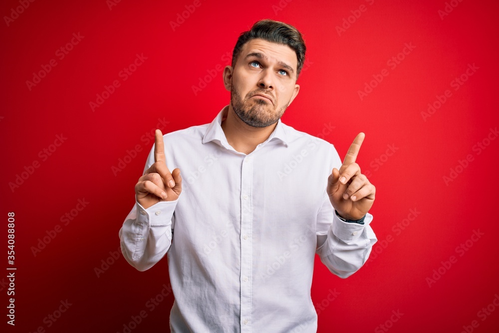 Young business man with blue eyes wearing elegant shirt standing over red isolated background Pointing up looking sad and upset, indicating direction with fingers, unhappy and depressed. Young business man with blue eyes wearing elegant shirt standing over red isolated background Pointing up looking sad and upset, indicating direction with fingers, unhappy and depressed.