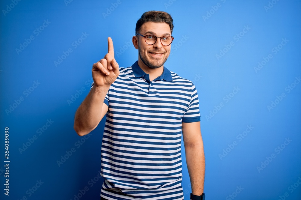 Young man with blue eyes wearing glasses and casual striped t-shirt over blue background showing and pointing up with finger number one while smiling confident and happy. Young man with blue eyes wearing glasses and casual striped t-shirt over blue background showing and pointing up with finger number one while smiling confident and happy.