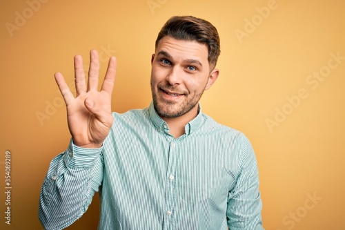 Young business man with blue eyes wearing elegant green shirt over yellow background showing and pointing up with fingers number four while smiling confident and happy.