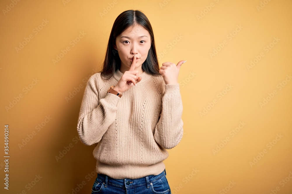 Fototapeta premium Young beautiful asian woman wearing casual sweater over yellow isolated background asking to be quiet with finger on lips pointing with hand to the side. Silence and secret concept.