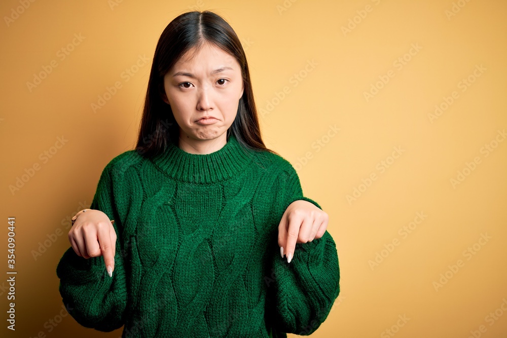Young beautiful asian woman wearing green winter sweater over yellow isolated background Pointing down looking sad and upset, indicating direction with fingers, unhappy and depressed. Young beautiful asian woman wearing green winter sweater over yellow isolated background Pointing down looking sad and upset, indicating direction with fingers, unhappy and depressed.