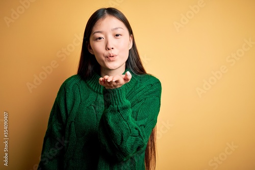 Young beautiful asian woman wearing green winter sweater over yellow isolated background looking at the camera blowing a kiss with hand on air being lovely and sexy. Love expression. © Krakenimages.com