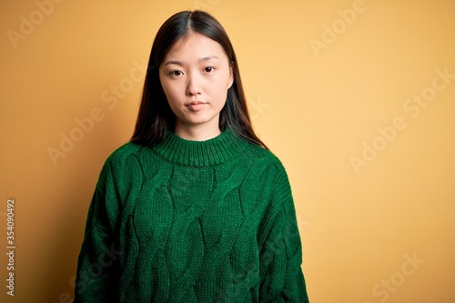 Young beautiful asian woman wearing green winter sweater over yellow isolated background Relaxed with serious expression on face. Simple and natural looking at the camera. © Krakenimages.com