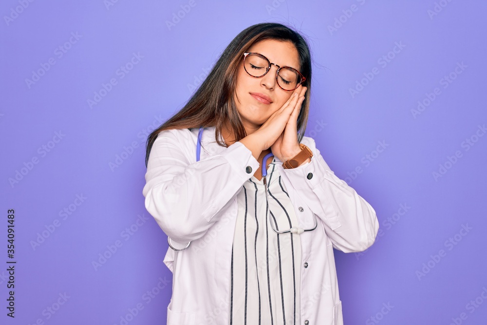 Professional doctor woman wearing stethoscope and medical coat over purple background sleeping tired dreaming and posing with hands together while smiling with closed eyes. Professional doctor woman wearing stethoscope and medical coat over purple background sleeping tired dreaming and posing with hands together while smiling with closed eyes.