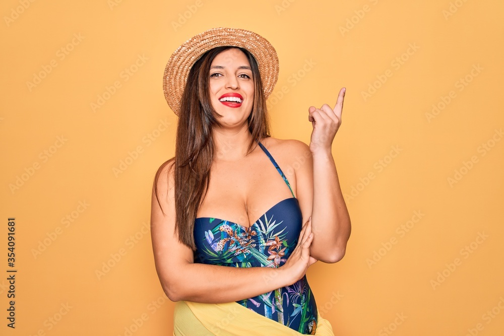 Young hispanic woman wearing summer hat and swimsuit over yellow background with a big smile on face, pointing with hand and finger to the side looking at the camera. Young hispanic woman wearing summer hat and swimsuit over yellow background with a big smile on face, pointing with hand and finger to the side looking at the camera.