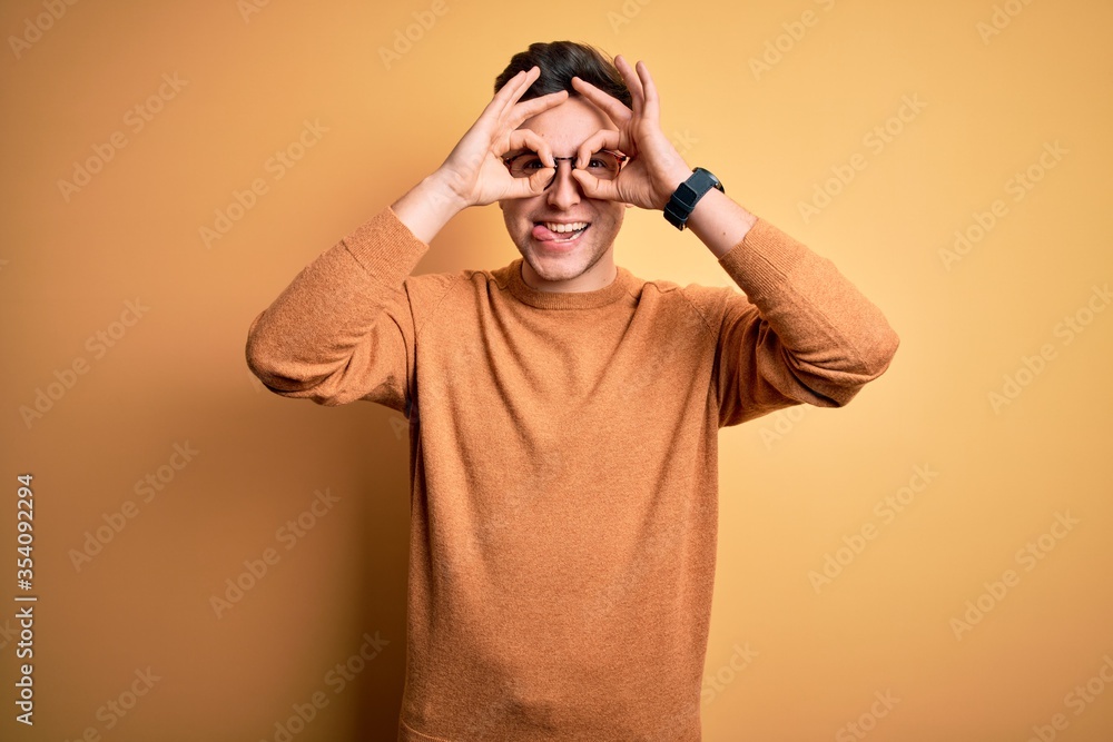 Young handsome caucasian man wearing glasses and casual winter sweater over yellow background doing ok gesture like binoculars sticking tongue out, eyes looking through fingers. Crazy expression. Young handsome caucasian man wearing glasses and casual winter sweater over yellow background doing ok gesture like binoculars sticking tongue out, eyes looking through fingers. Crazy expression.