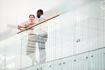 Low angle view at two contemporary business people chatting cheerfully while standing on balcony in minimal office building interior, copy space
