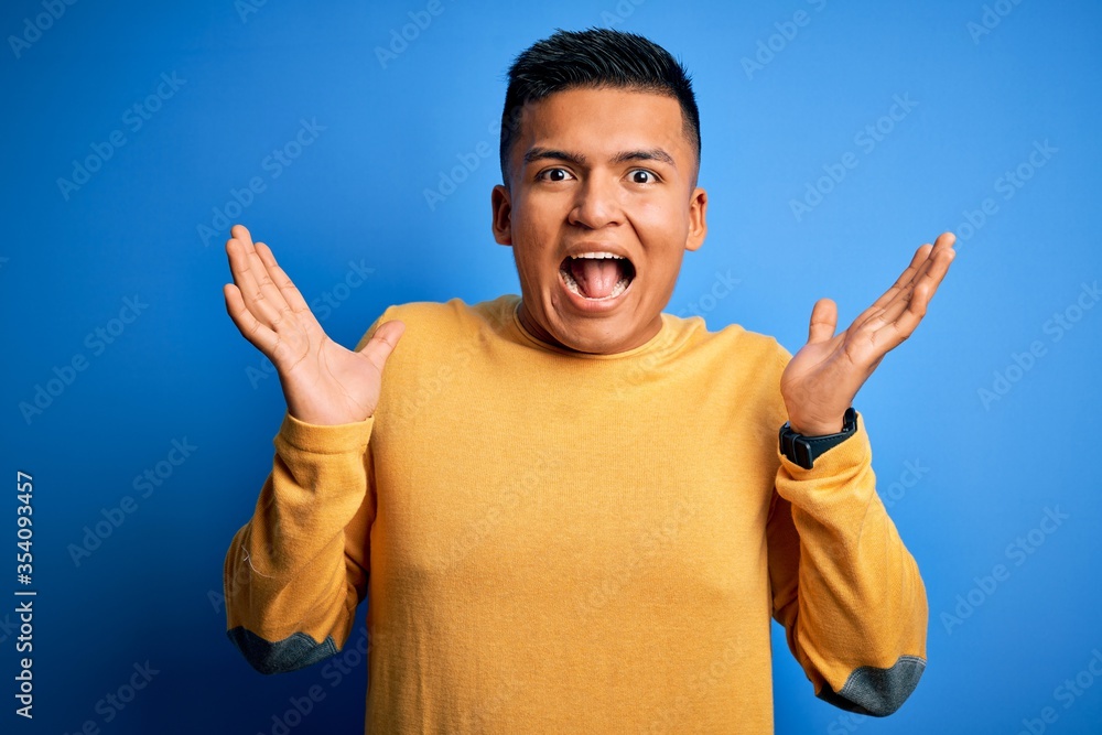 Young handsome latin man wearing yellow casual sweater over isolated blue background celebrating crazy and amazed for success with arms raised and open eyes screaming excited. Winner concept Young handsome latin man wearing yellow casual sweater over isolated blue background celebrating crazy and amazed for success with arms raised and open eyes screaming excited. Winner concept