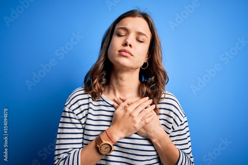 Young beautiful blonde girl wearing casual sweater standing over blue isolated background smiling with hands on chest with closed eyes and grateful gesture on face. Health concept.