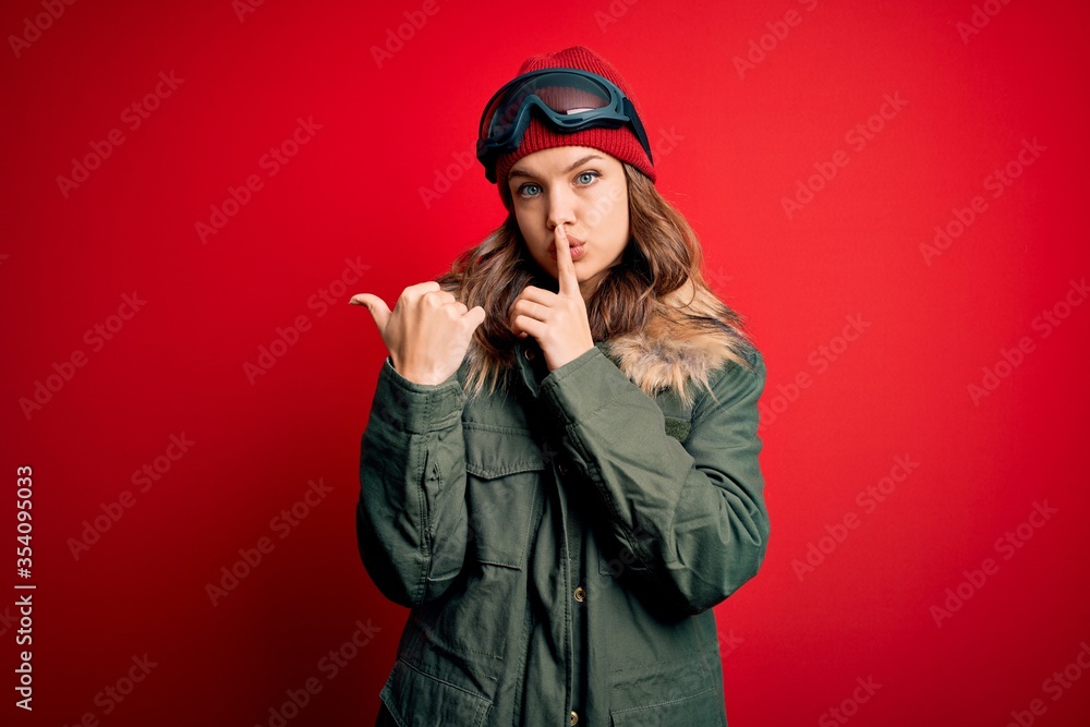 Young blonde girl wearing ski glasses and winter coat for ski weather over red background asking to be quiet with finger on lips pointing with hand to the side. Silence and secret concept.