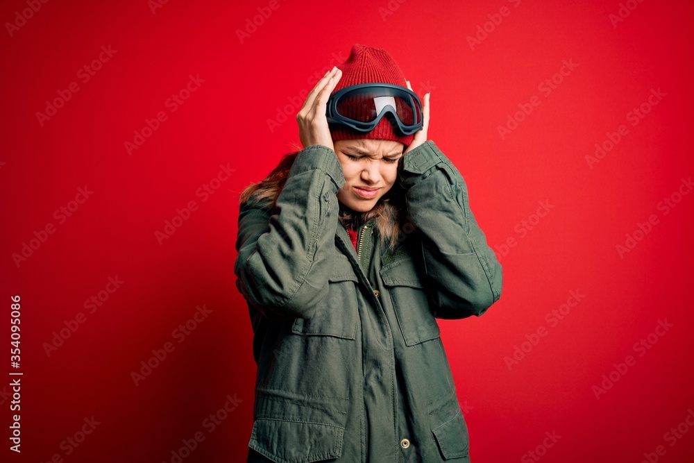 Young blonde girl wearing ski glasses and winter coat for ski weather over red background suffering from headache desperate and stressed because pain and migraine. Hands on head.