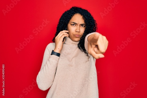 Young african american curly woman having conversation talking on the smartphone pointing with finger to the camera and to you  hand sign  positive and confident gesture from the front