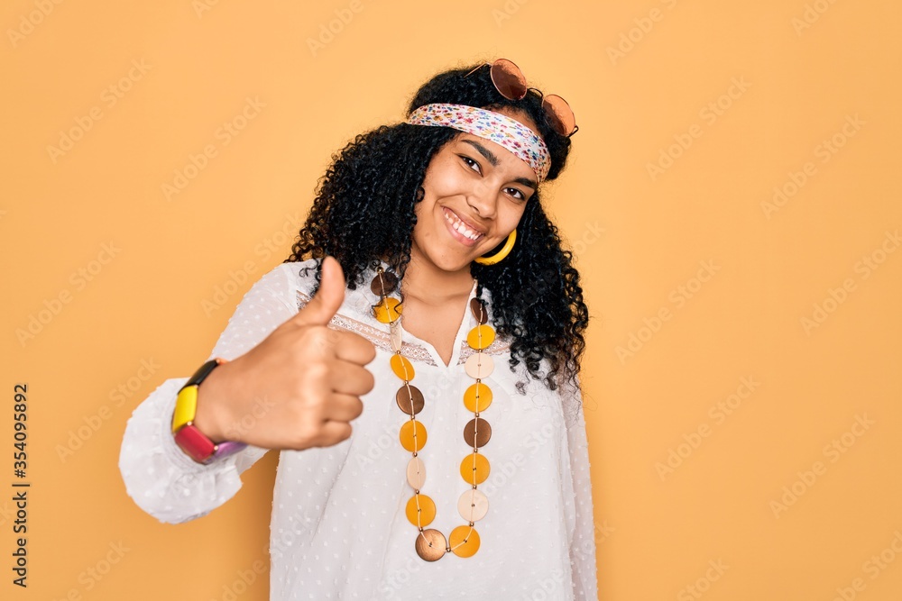 Young african american curly hippie woman wearing sunglasses and vintage accessories doing happy thumbs up gesture with hand. Approving expression looking at the camera showing success.