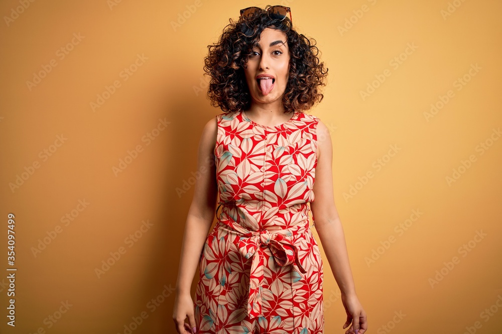 Young beautiful curly arab woman on vacation wearing summer floral dress and sunglasses sticking tongue out happy with funny expression. Emotion concept. Young beautiful curly arab woman on vacation wearing summer floral dress and sunglasses sticking tongue out happy with funny expression. Emotion concept.