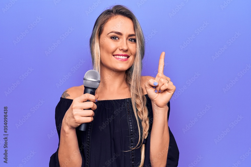 Young beautiful blonde singer woman singing using microphone over purple background smiling with an idea or question pointing finger with happy face, number one Young beautiful blonde singer woman singing using microphone over purple background smiling with an idea or question pointing finger with happy face, number one