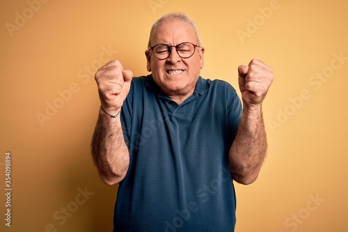 Middle age handsome hoary man wearing casual polo and glasses over yellow background excited for success with arms raised and eyes closed celebrating victory smiling. Winner concept.