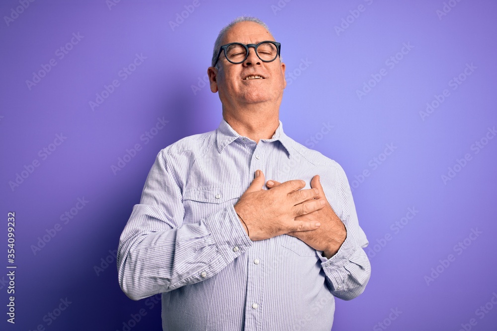 Middle age handsome hoary man wearing striped shirt and glasses over purple background smiling with hands on chest with closed eyes and grateful gesture on face. Health concept. Middle age handsome hoary man wearing striped shirt and glasses over purple background smiling with hands on chest with closed eyes and grateful gesture on face. Health concept.