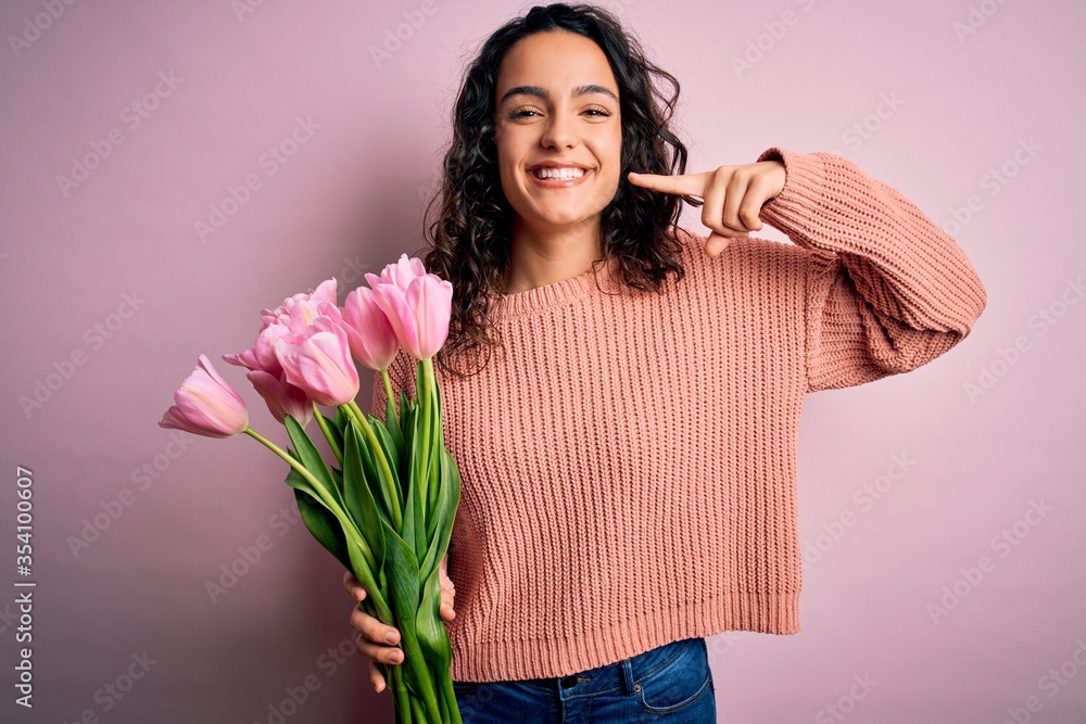 Young beautiful romantic woman with curly hair holding bouquet of pink tulips smiling cheerful showing and pointing with fingers teeth and mouth. Dental health concept. Young beautiful romantic woman with curly hair holding bouquet of pink tulips smiling cheerful showing and pointing with fingers teeth and mouth. Dental health concept.