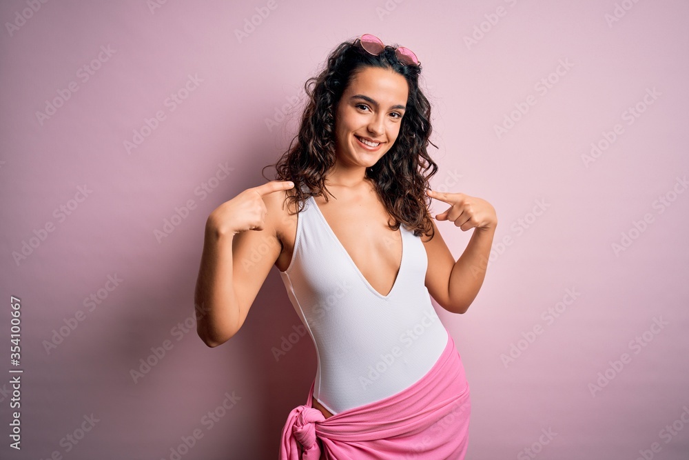 Beautiful woman with curly hair on vacation wearing white swimsuit over pink background looking confident with smile on face, pointing oneself with fingers proud and happy. Beautiful woman with curly hair on vacation wearing white swimsuit over pink background looking confident with smile on face, pointing oneself with fingers proud and happy.