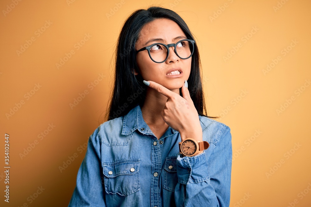 Young beautiful chinese woman wearing casual denim shirt over isolated yellow background Thinking worried about a question, concerned and nervous with hand on chin