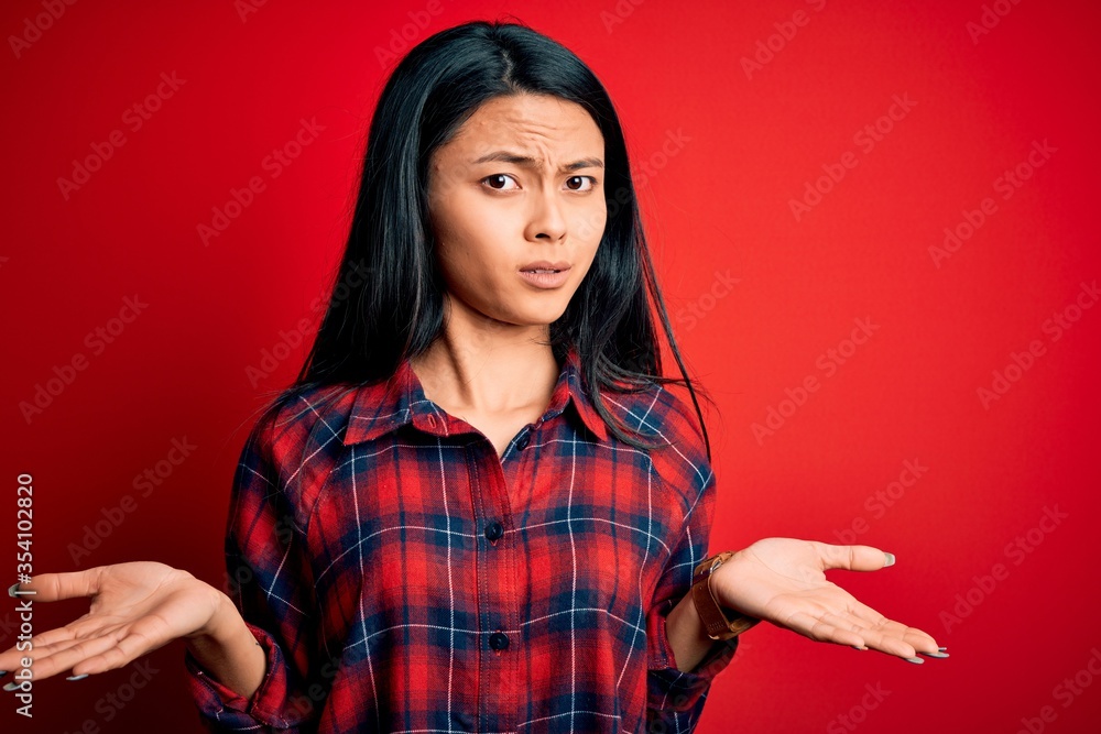Young beautiful chinese woman wearing casual shirt over isolated red background clueless and confused with open arms, no idea concept. Young beautiful chinese woman wearing casual shirt over isolated red background clueless and confused with open arms, no idea concept.