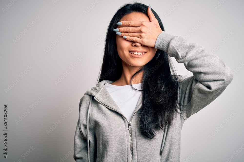 Young beautiful chinese sporty woman wearing sweatshirt over isolated white background smiling and laughing with hand on face covering eyes for surprise. Blind concept. Young beautiful chinese sporty woman wearing sweatshirt over isolated white background smiling and laughing with hand on face covering eyes for surprise. Blind concept.