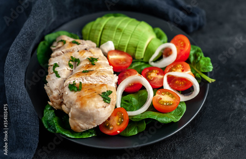  Grilled chicken breast and avocado salad with cherry tomatoes, spinach, in a black plate on a stone background