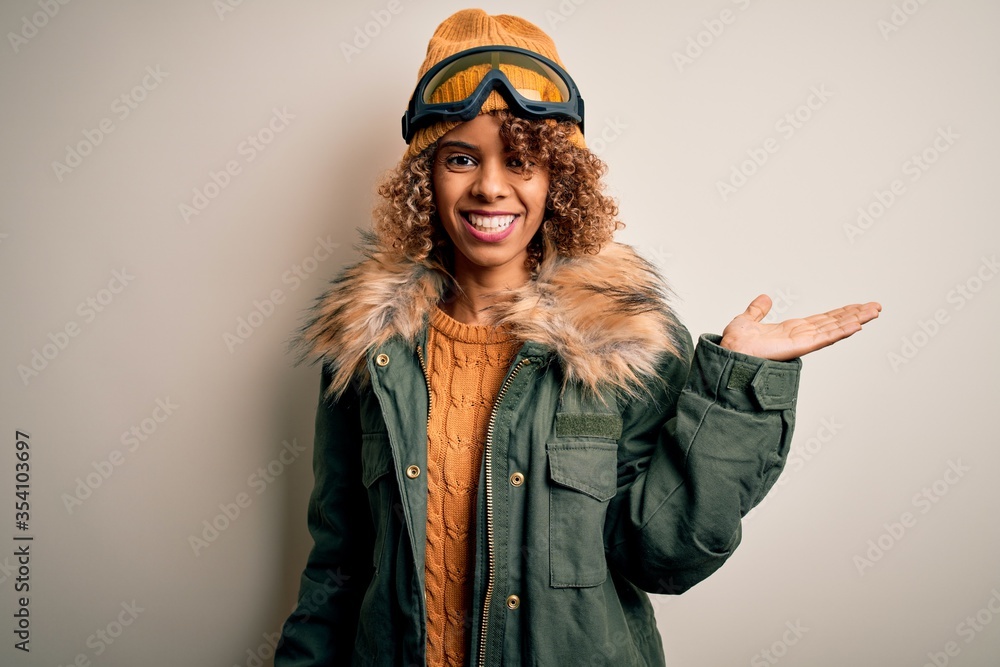 Young african american skier woman with curly hair wearing snow sportswear and ski goggles smiling cheerful presenting and pointing with palm of hand looking at the camera. Young african american skier woman with curly hair wearing snow sportswear and ski goggles smiling cheerful presenting and pointing with palm of hand looking at the camera.