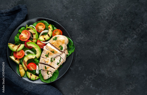  Grilled chicken breast and grilled avocado salad with cherry tomatoes, spinach, in a black plate on a stone background with copy space for your text