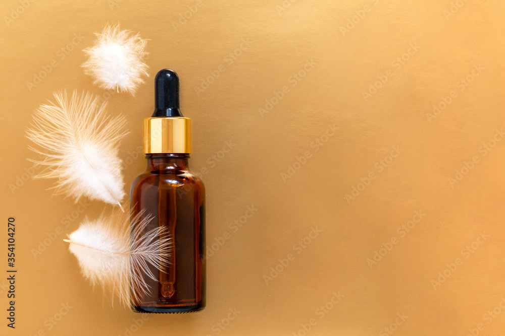 Fototapeta premium Oil bottle with pipette on the golden background with white feathers and copy space. Skin care concept