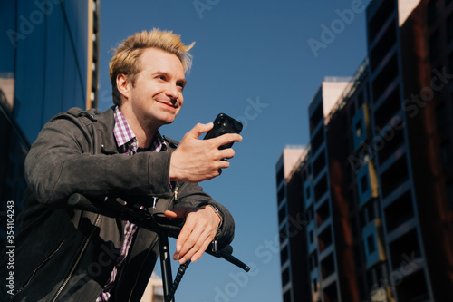 A man in everyday clothes stands at the scooter and look into the smartphone