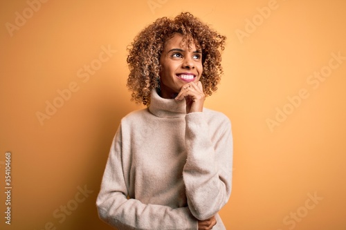 Young beautiful african american woman wearing turtleneck sweater over yellow background with hand on chin thinking about question, pensive expression. Smiling and thoughtful face. Doubt concept.