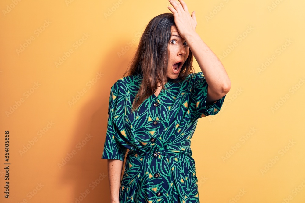 Young beautiful brunette woman wearing casual floral dress standing over yellow background surprised with hand on head for mistake, remember error. Forgot, bad memory concept. Young beautiful brunette woman wearing casual floral dress standing over yellow background surprised with hand on head for mistake, remember error. Forgot, bad memory concept.