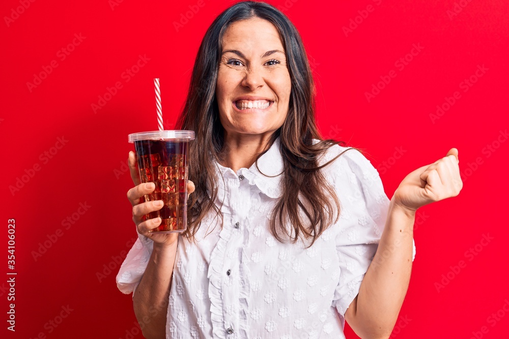 Beautiful brunette woman drinking cola refreshment beverage using straw over red background screaming proud, celebrating victory and success very excited with raised arm Beautiful brunette woman drinking cola refreshment beverage using straw over red background screaming proud, celebrating victory and success very excited with raised arm