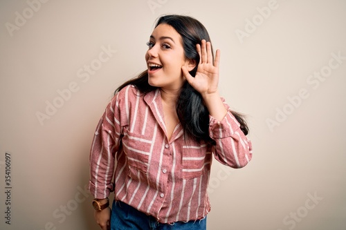 Young brunette woman wearing casual striped shirt over isolated background smiling with hand over ear listening an hearing to rumor or gossip. Deafness concept.
