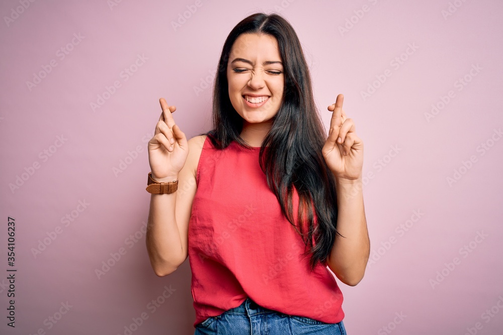 Young brunette woman wearing casual summer shirt over pink isolated background gesturing finger crossed smiling with hope and eyes closed. Luck and superstitious concept. Young brunette woman wearing casual summer shirt over pink isolated background gesturing finger crossed smiling with hope and eyes closed. Luck and superstitious concept.