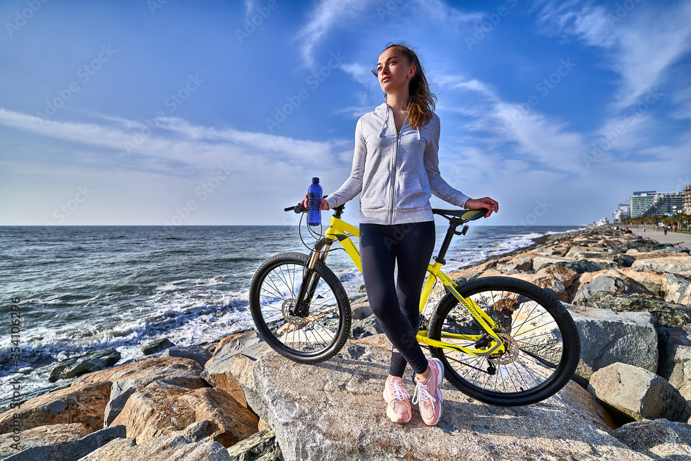 Fototapeta premium Fitness woman with yellow bicycle stands on the stones on the seashore in a clear sunny day against the blue sky. Athletic healthy people with active and sporty lifestyle