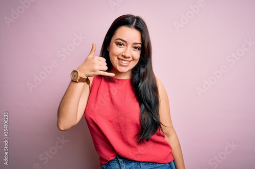 Young brunette woman wearing casual summer shirt over pink isolated background smiling doing phone gesture with hand and fingers like talking on the telephone. Communicating concepts.