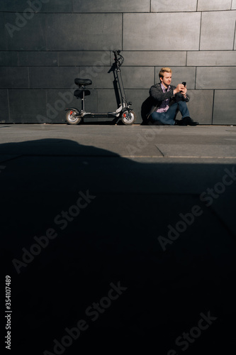 Young male freelancer with smartphone and electric scooter sitting on the road near a modern business center