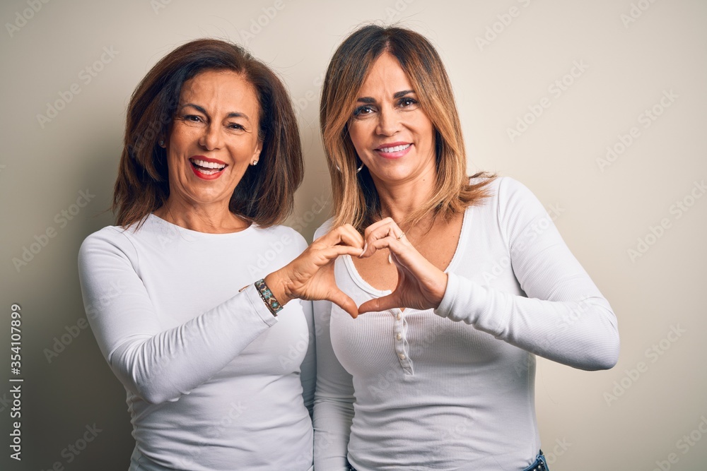Middle age beautiful couple of sisters wearing casual t-shirt over isolated white background smiling in love showing heart symbol and shape with hands. Romantic concept. Middle age beautiful couple of sisters wearing casual t-shirt over isolated white background smiling in love showing heart symbol and shape with hands. Romantic concept.