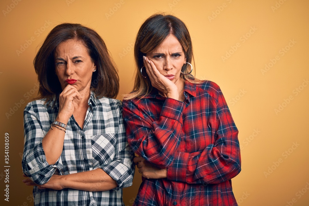 Middle age beautiful couple of sisters wearing casual shirt over isolated yellow background thinking looking tired and bored with depression problems with crossed arms. Middle age beautiful couple of sisters wearing casual shirt over isolated yellow background thinking looking tired and bored with depression problems with crossed arms.