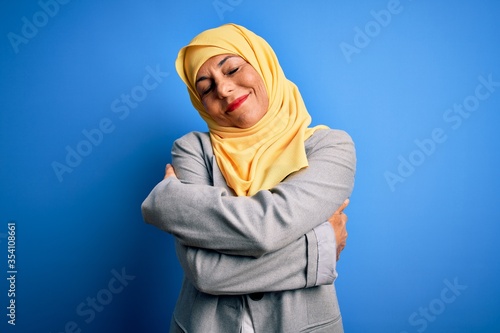 Middle age brunette business woman wearing muslim traditional hijab over blue background Hugging oneself happy and positive, smiling confident. Self love and self care © Krakenimages.com
