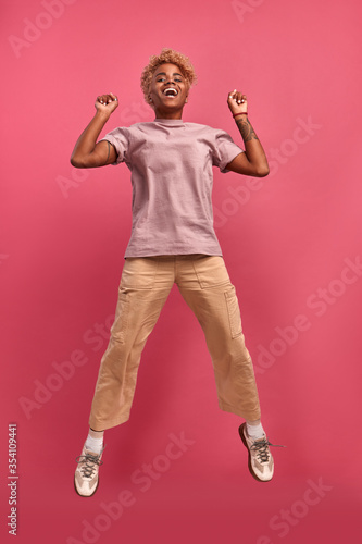 Full length body shot of emotional african american student female jumps happily in air, feels energetic and optimistic on pink studio background. Concept of services for youth in the banking sector