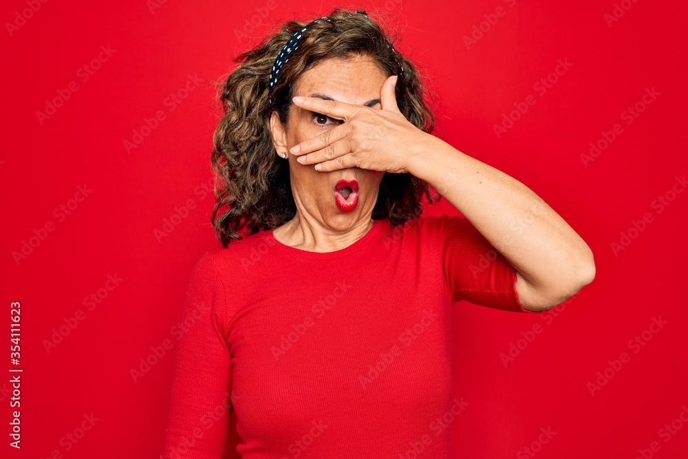Middle age senior brunette woman wearing casual t-shirt standing over red background peeking in shock covering face and eyes with hand, looking through fingers with embarrassed expression. Middle age senior brunette woman wearing casual t-shirt standing over red background peeking in shock covering face and eyes with hand, looking through fingers with embarrassed expression.