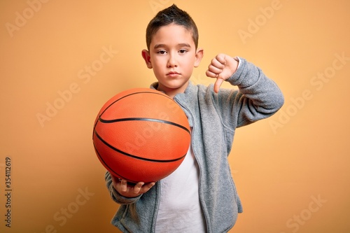Young little boy kid playing with basketball game ball over isolated yellow background with angry face, negative sign showing dislike with thumbs down, rejection concept © Krakenimages.com