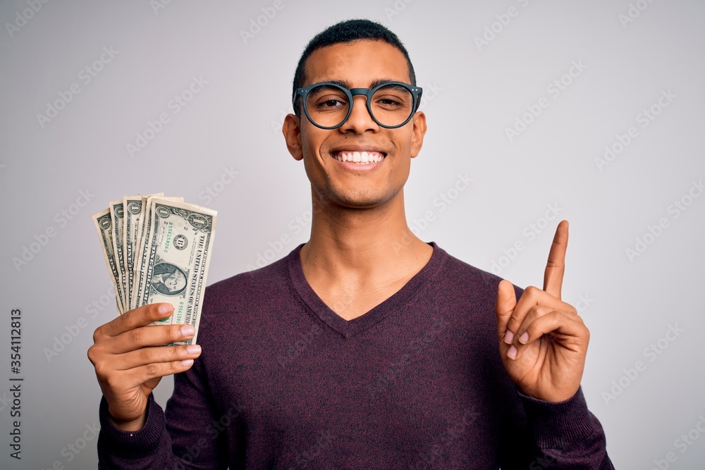 Handsome african american man holding bunch of dollars banknotes over white background surprised with an idea or question pointing finger with happy face, number one Handsome african american man holding bunch of dollars banknotes over white background surprised with an idea or question pointing finger with happy face, number one